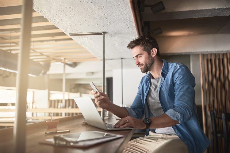 man sitting a table with his laptop looking at this phone 