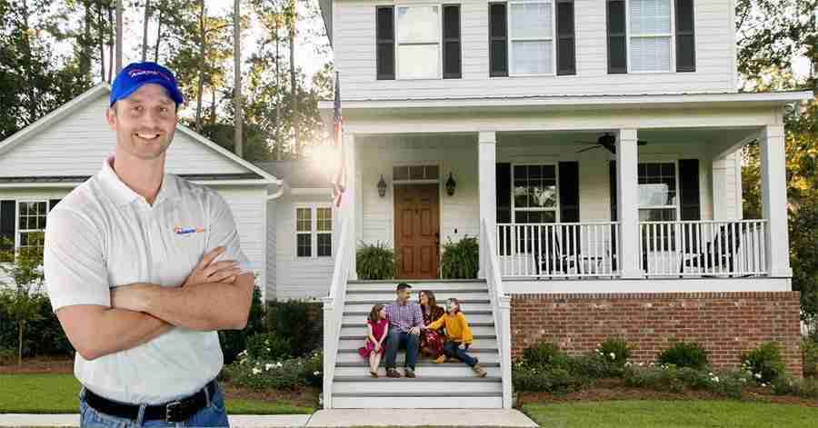 A man with his arms crossed standing in front of a house where a family is sitting on the steps. 