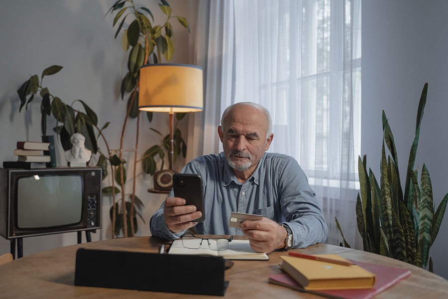 An older man sitting at a desk holding a phone and credit card.