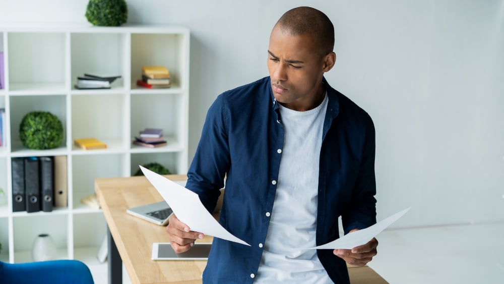 A person leaned up against a desk comparing two pieces of paper. 