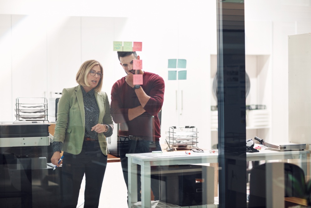 Two people observing post-it notes on a glass window. 