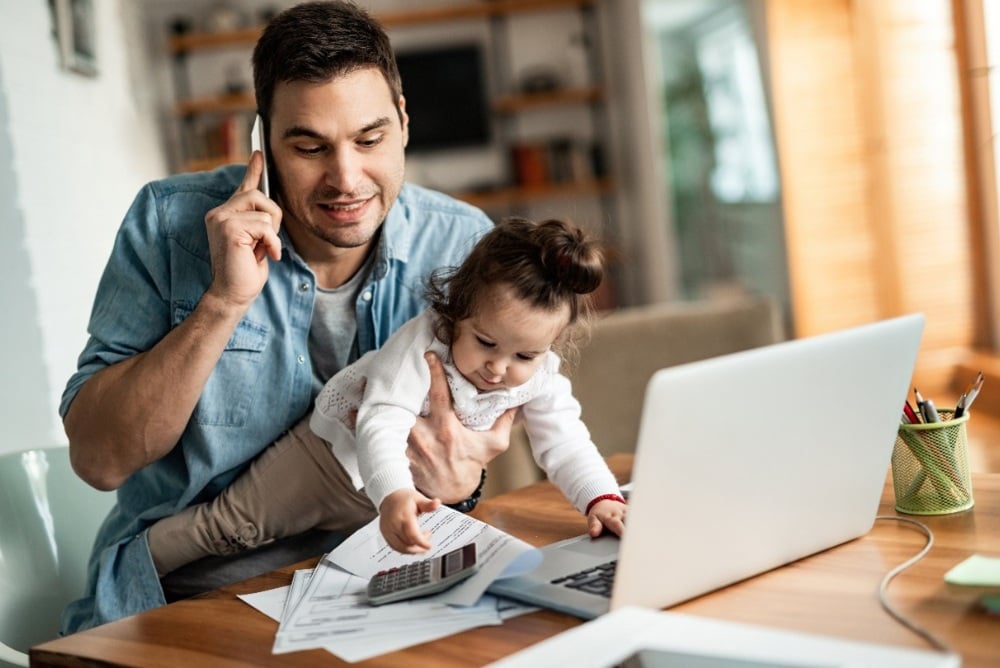 A man holding a baby in front of a laptop, papers, and a calculator. 